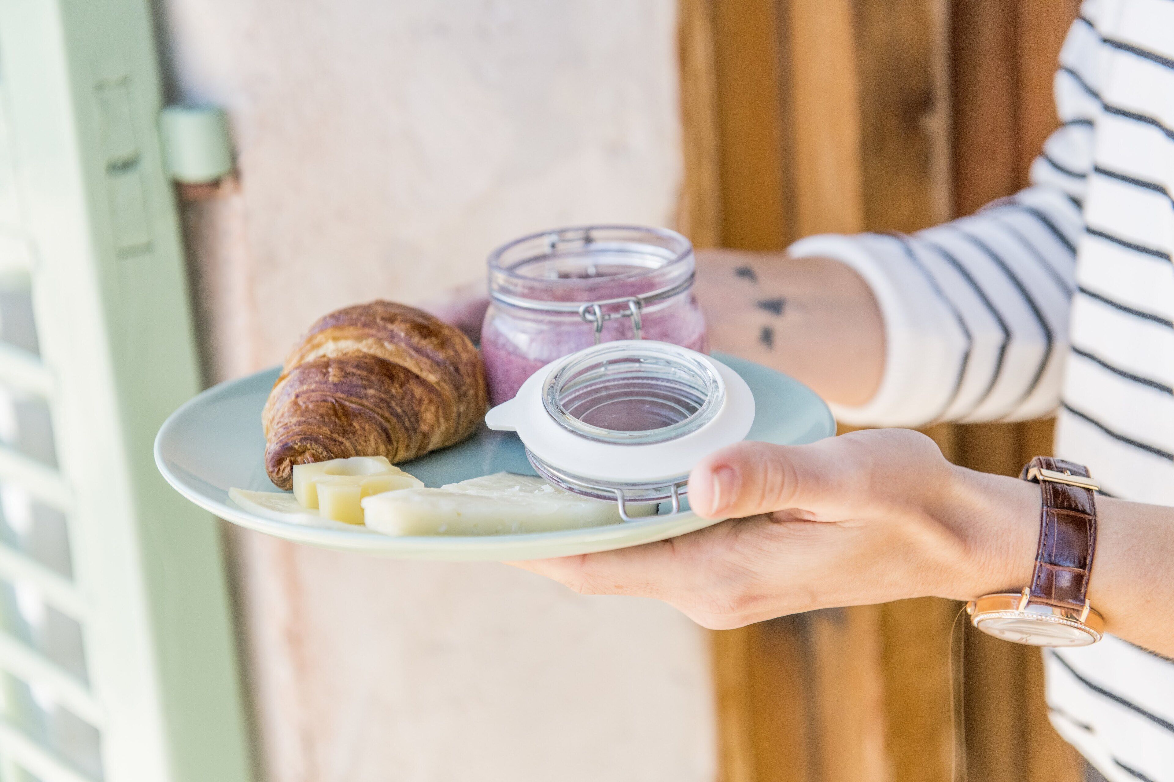 Una persona sosteniendo un plato con un croissant, queso y un frasco de mermelada.