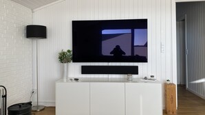 Una sala de estar moderna con un televisor de pantalla plana montado en una pared blanca, una lámpara de pie, una consola blanca con adornos y un piso de madera.