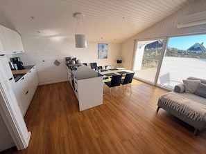 A modern kitchen with a central island, a dining area with a table and chairs, and a large sliding glass door leading to a balcony. - Well-equipped summer house with a charging port for electric cars! (Hvide Sande)