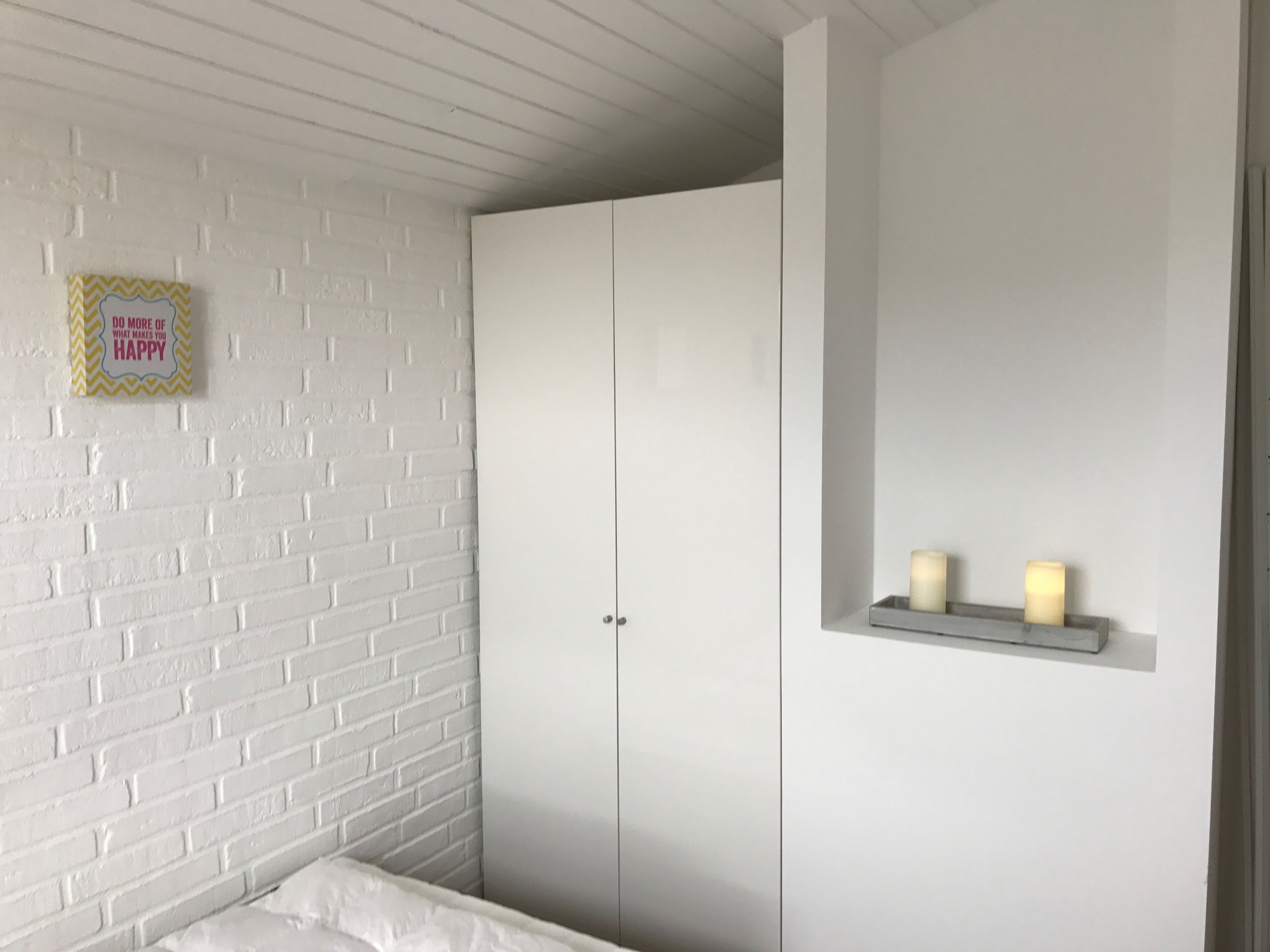 Ett sovrum med vita tegelväggar, en vit garderob och en hylla vid sängen med ljus.