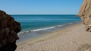 Une plage avec une eau d’un bleu limpide, du sable composé de petits galets et des falaises rocheuses.