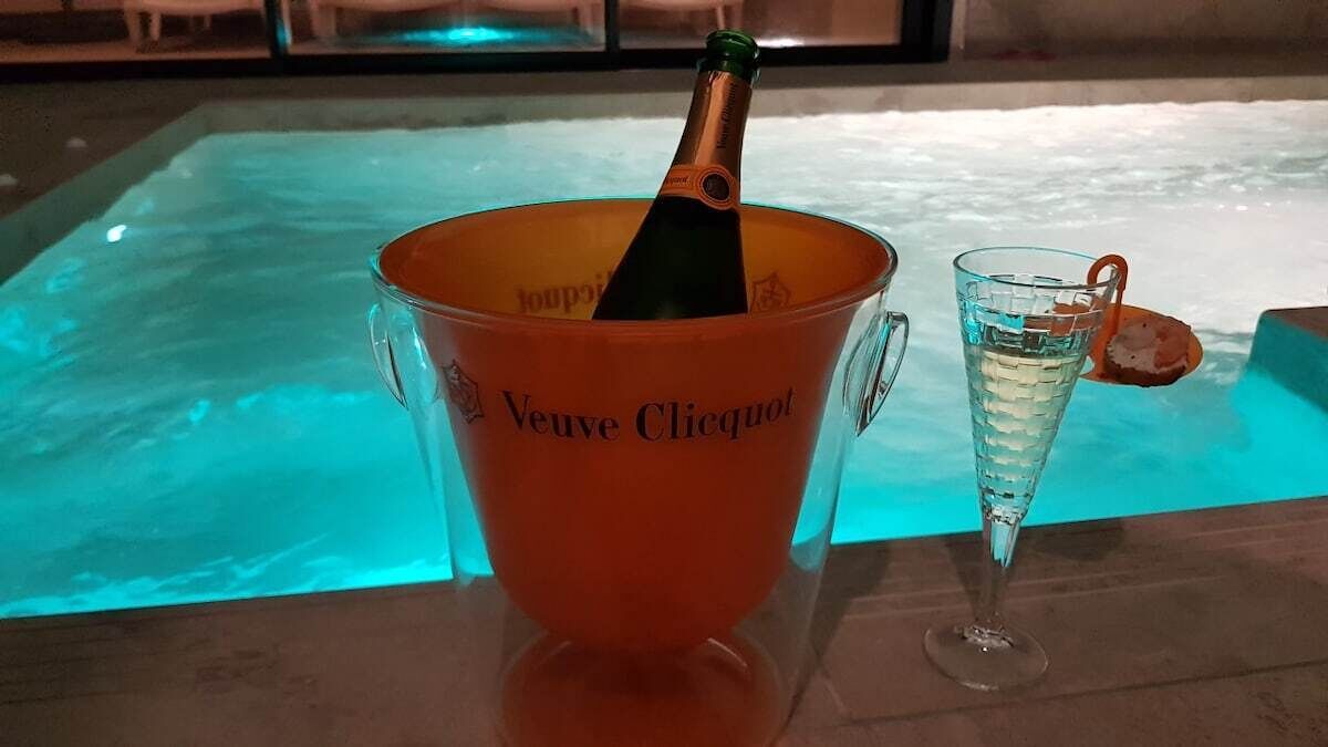 Une bouteille de champagne Veuve Clicquot dans un seau à glaçons de la même marque, à côté d’une coupe à champagne, sur une table au bord de la piscine.