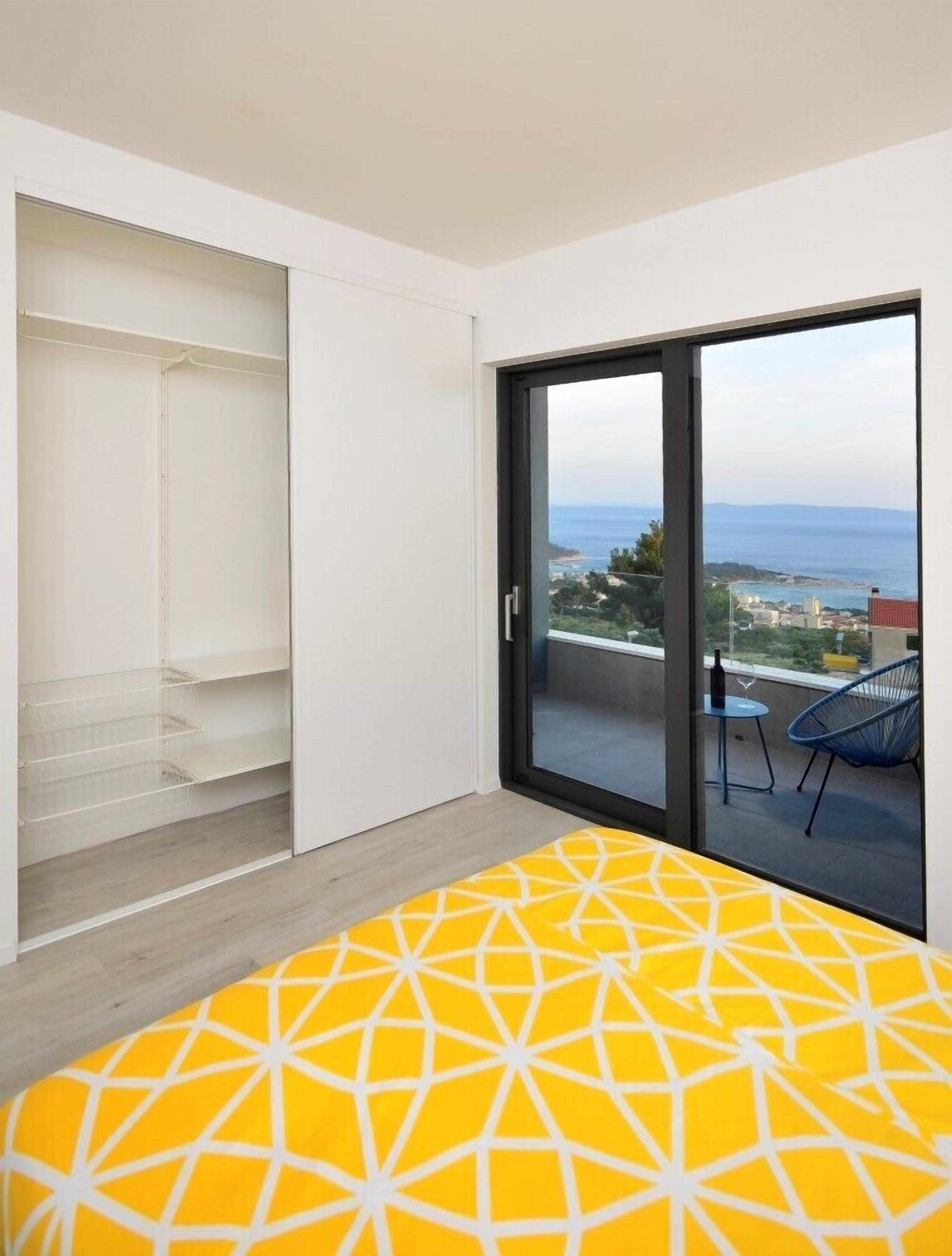 Una habitación moderna con una colcha amarilla estampada, una puerta corrediza de vidrio que da a un balcón con vista al mar y un armario blanco con estantes abiertos.