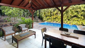 Um alpendre coberto com móveis de madeira, piscina e vegetação exuberante.