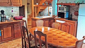Una cocina con armarios de madera, una mesa de comedor y sillas. Hay un microondas, un refrigerador y un lavabo. Se ve una escalera al fondo.
