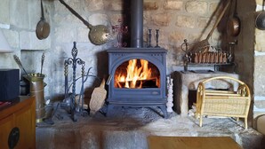 A stone fireplace with a black stove, surrounded by various antique kitchen utensils and a wicker chair.
