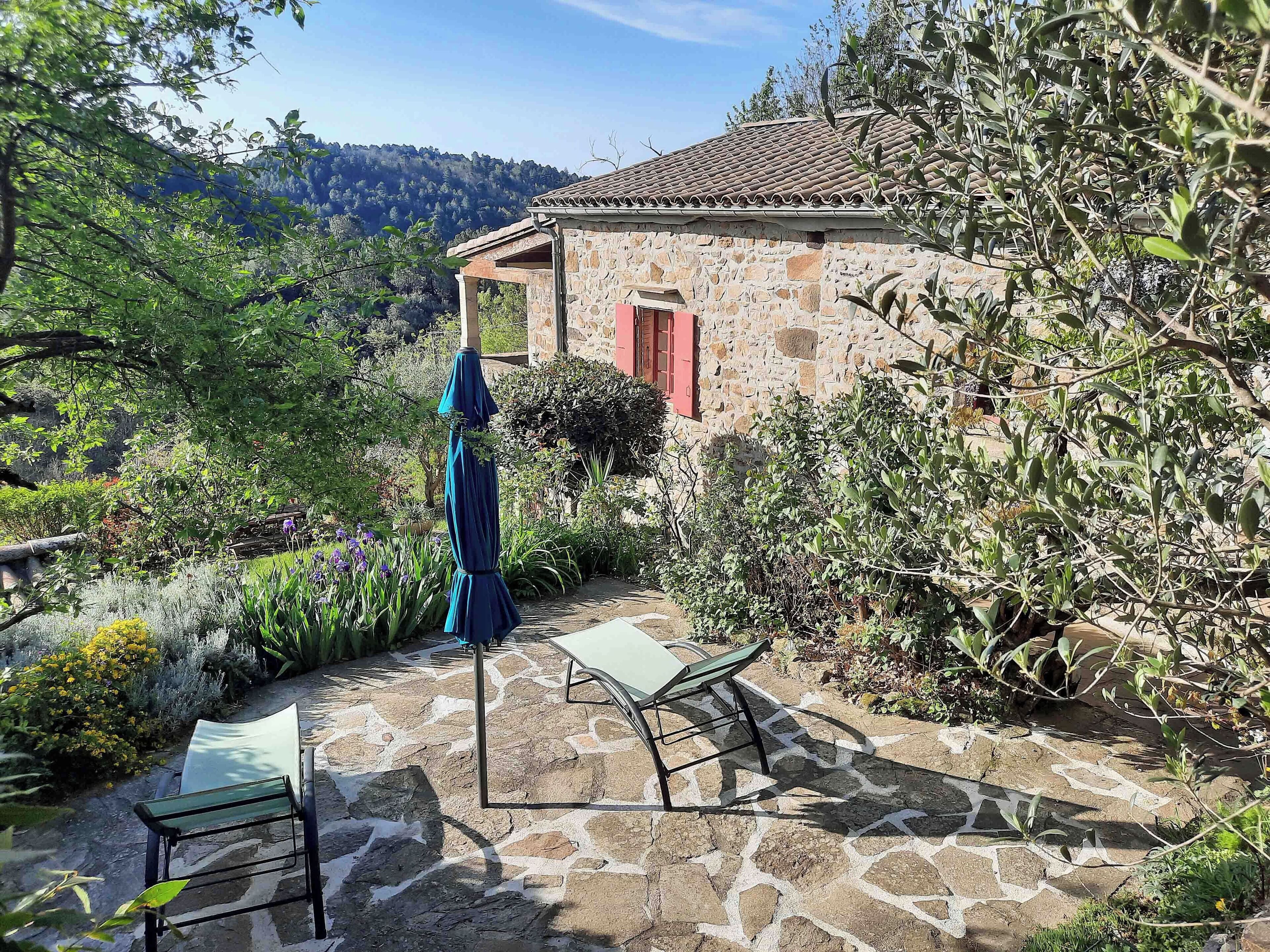 A stone building with a tiled roof, surrounded by lush greenery and a stone patio with a table and chairs.