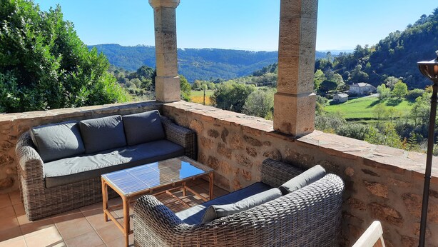 A covered outdoor seating area with wicker furniture, a glass-top table, and a view of a green valley.