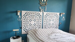 A bedroom with a teal wall, a white bed with a striped bedspread, a decorative headboard, and a small bedside table with a lamp.