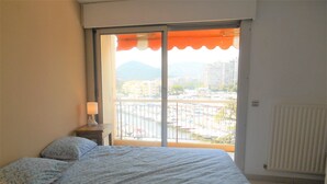 Ein Schlafzimmer mit Bett, Nachttisch, Fenster mit orangefarbener Markise und Blick auf einen Yachthafen und Gebäude.