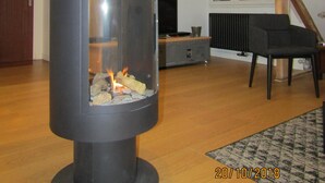 Un soggiorno moderno con una stufa a legna in legno nero, travi in legno, una televisione e una poltrona nera.