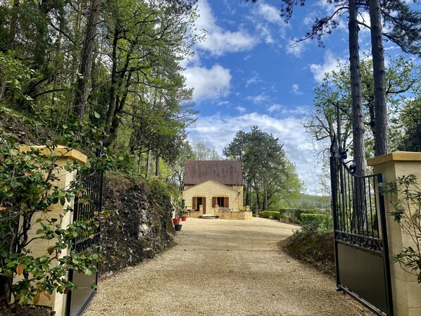 Um edifício de pedra com uma entrada portão, acessível por um caminho de cascalho, cercado por árvores e um céu limpo.