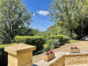 Uma trilha de pedra serpenteia por um jardim exuberante, com sebes bem aparadas e vista para uma colina com uma construção semelhante a um castelo ao longe.
