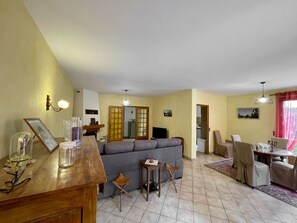 Sala de estar com sofá, mesa de jantar com cadeiras, televisão e cozinha visível através de uma porta aberta.