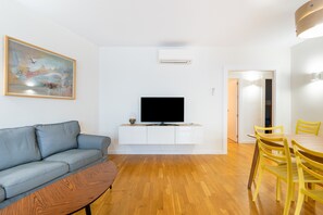 A modern living room with a grey sofa, a flat-screen TV on a white cabinet, wooden flooring, and a dining area with yellow chairs.