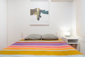A room with a bed, striped bedding, two pillows, a wall-mounted abstract painting, a bedside table with a lamp, and a white nightstand.