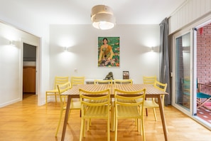 A dining area with a wooden table and yellow chairs, a painting on the wall, and a sliding glass door to the right.
