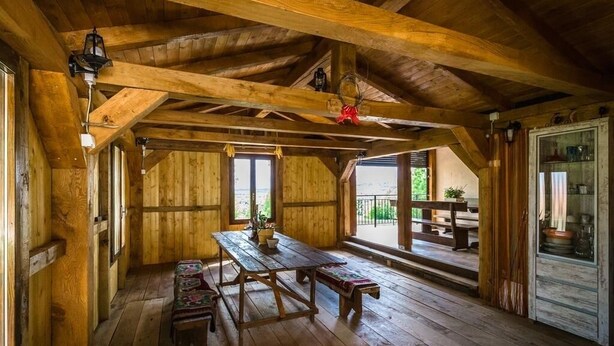 A wooden interior with a high ceiling, wooden beams, a central table with benches, and large windows.