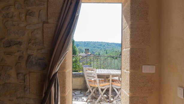 Une pièce avec un mur en pierre, une fenêtre avec des rideaux, une table et des chaises en bois, et une vue sur un paysage verdoyant.