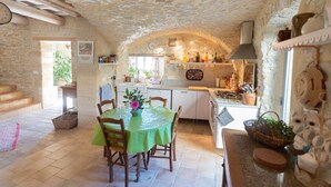 Une cuisine rustique aux murs de pierre, un coin repas avec une table dressée pour deux personnes, et une vue sur l’extérieur par une fenêtre.