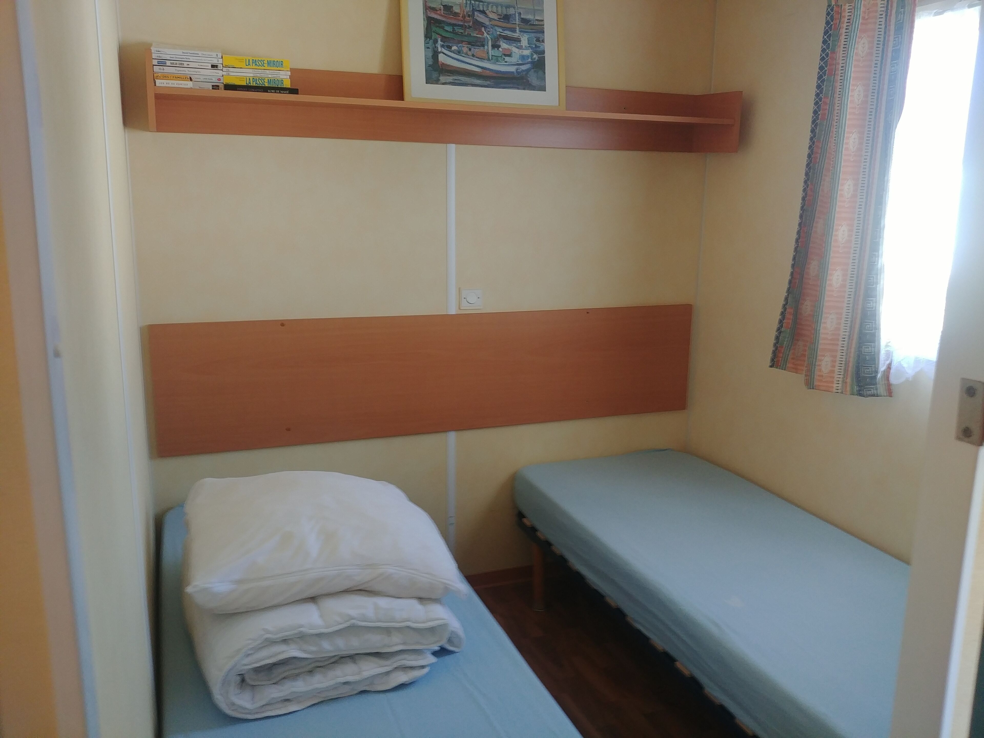 A small, single-bed room with a wooden headboard, a shelf with books, and a framed picture on the wall.