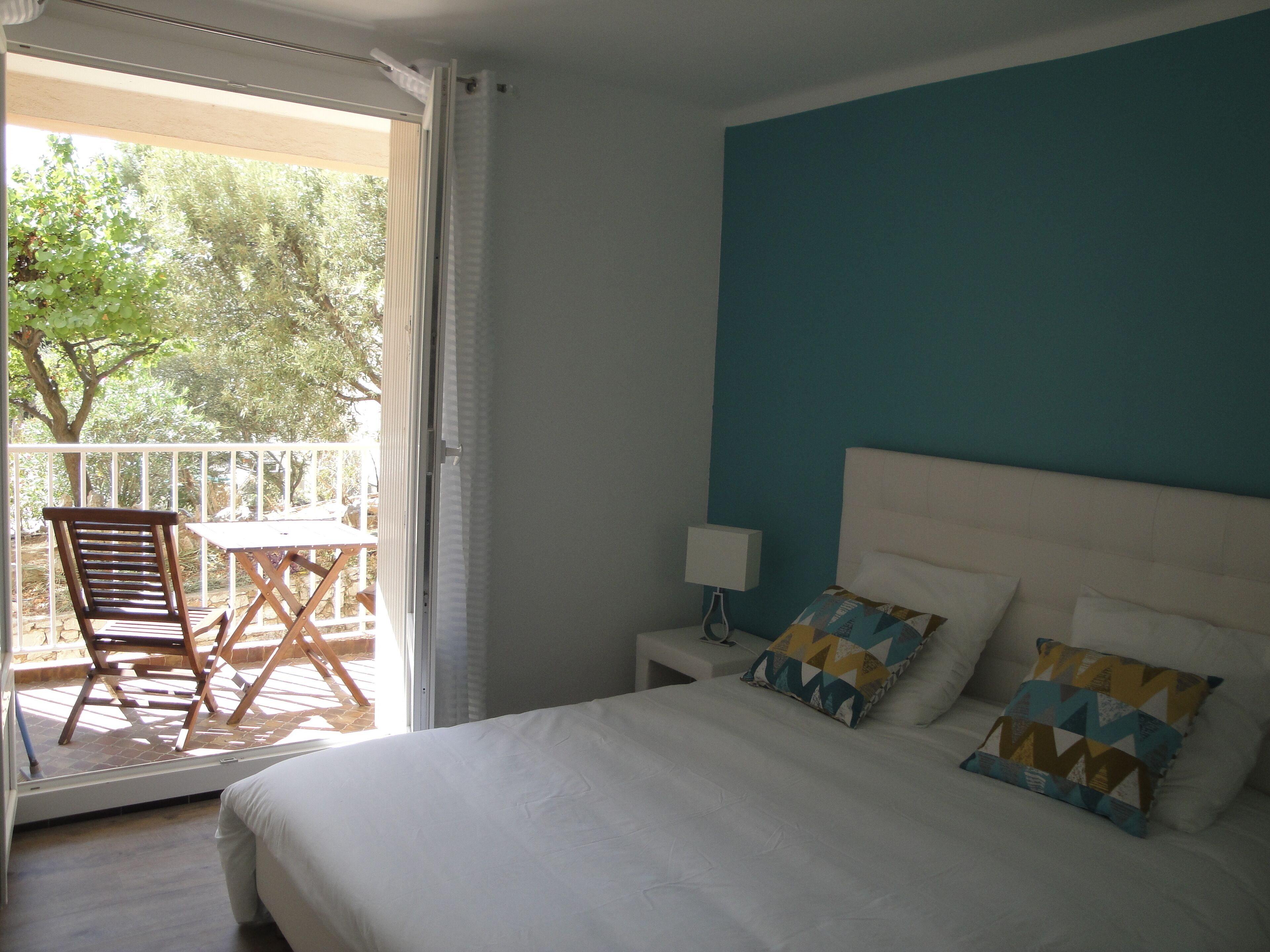 A bedroom with a bed, a nightstand, a lamp, a chair, and a view of the outdoors.