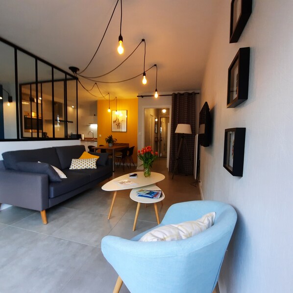 A modern living room with a sofa, a blue armchair, a coffee table, and a dining area in the background. - Apartment 4 people - Sea view (Bénodet)