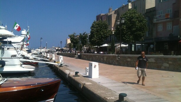 Un port esportiu amb vaixells amarrats, un home caminant per la vorera i edificis al fons.