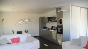 Un saló modern amb dos sofàs blancs, una zona de cuina amb electrodomèstics d'acer inoxidable i una lluminària de sostre singular.