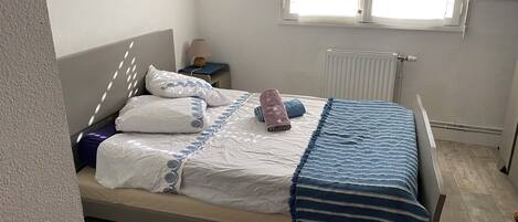 A neatly made bed with patterned pillows and a striped blanket, a bedside table with a lamp, and a radiator.
