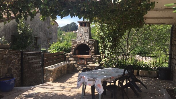 Eine Terrasss mit Steinofen, einem Tisch mit Tischdecke und Blick auf ein Steinhaus im Hintergrund.
