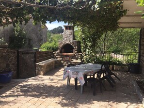 Eine Terrasss mit Steinofen, einem Tisch mit Tischdecke und Blick auf ein Steinhaus im Hintergrund.
