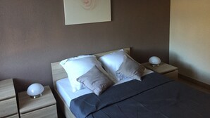 Ein ordentlich bezogenes Bett mit Kissen und Decke, ein Nachttisch mit Lampe sowie ein Wandbild.