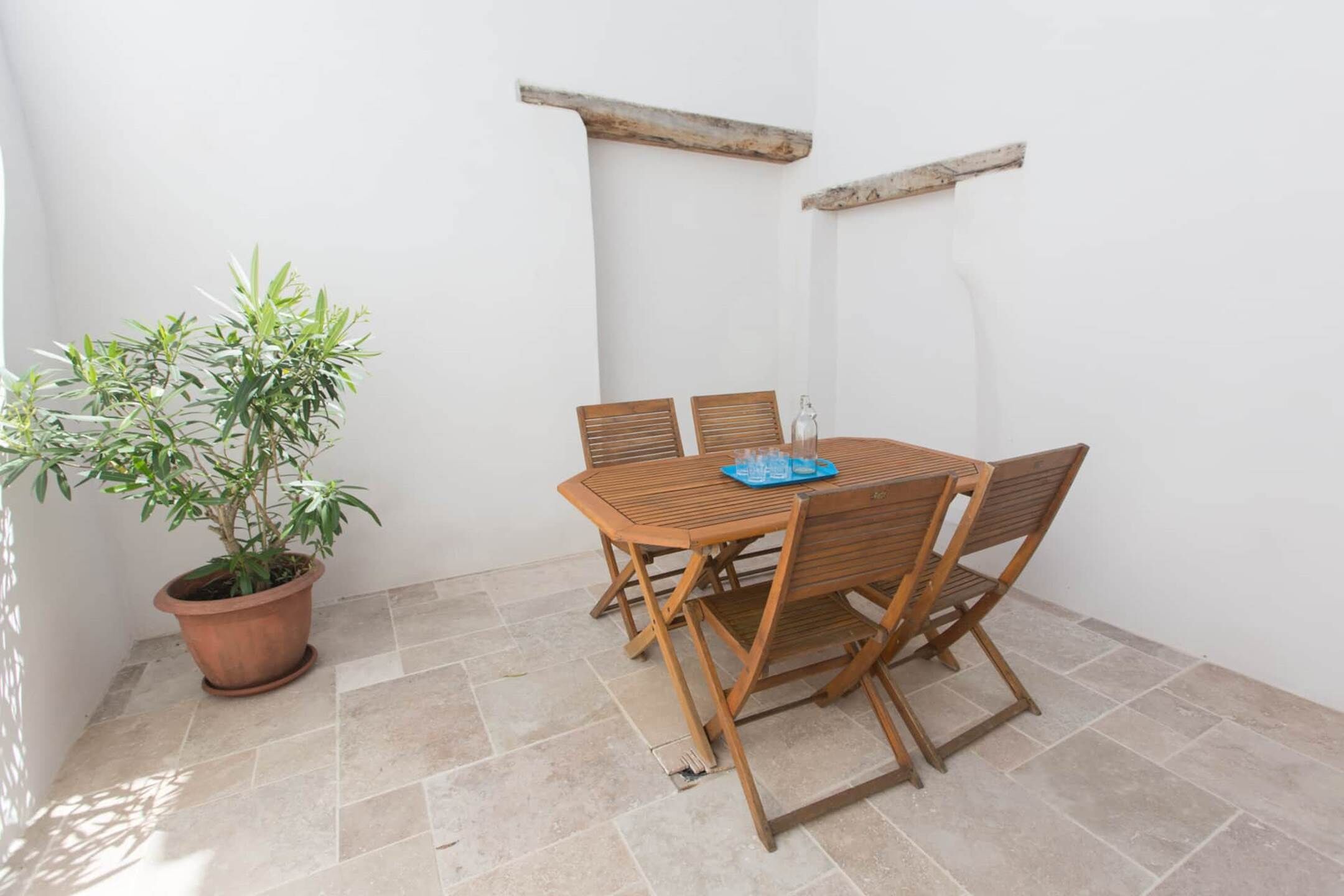 A small patio with a wooden table and chairs, a potted plant, and a white wall with a wooden shelf.