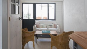 A room with a white sofa, two wicker chairs, a wooden table, and a window with blinds. - La Petite Maison (sleeps 4) (La Couarde-sur-Mer)