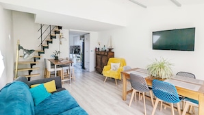 Uma sala de estar moderna com um sofá, mesa de jantar e cadeiras. Há uma escada que leva a um andar superior, e uma TV fixada na parede.