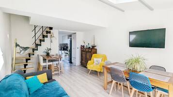 Un salon moderne avec un canapé, une table à manger et des chaises. Un escalier mène à l’étage supérieur, et un téléviseur est fixé au mur.
