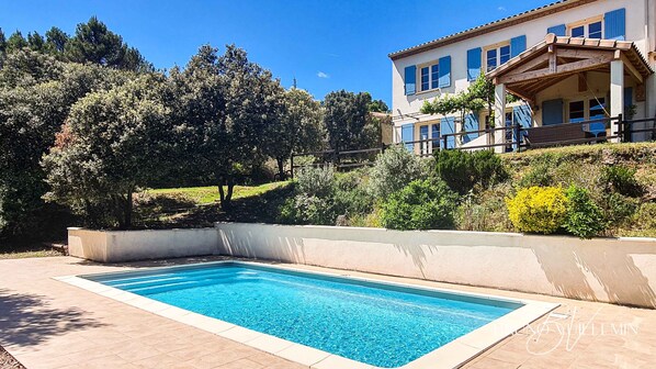 A swimming pool with a tiled deck, surrounded by a landscaped garden and a building with blue shutters. - Beautiful Provencal house (Caunes-Minervois)
