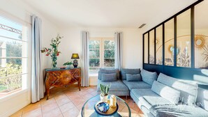 Ein Wohnzimmer mit einer Couch, einem Glastisch, einem Beistelltisch mit Lampe und Blick auf Grünflächen.