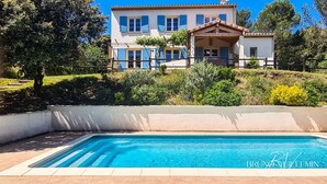 Ein Swimmingpool mit glatter, blauer Wasseroberfläche, umgeben von einer gefliesten Liegefläche. Im Hintergrund ein großes, zweistöckiges Haus mit blauen Fensterläden und einer überdachten Veranda.