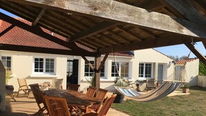 A wooden deck with a table and chairs, a hammock, and a house with a red-tiled roof in the background.