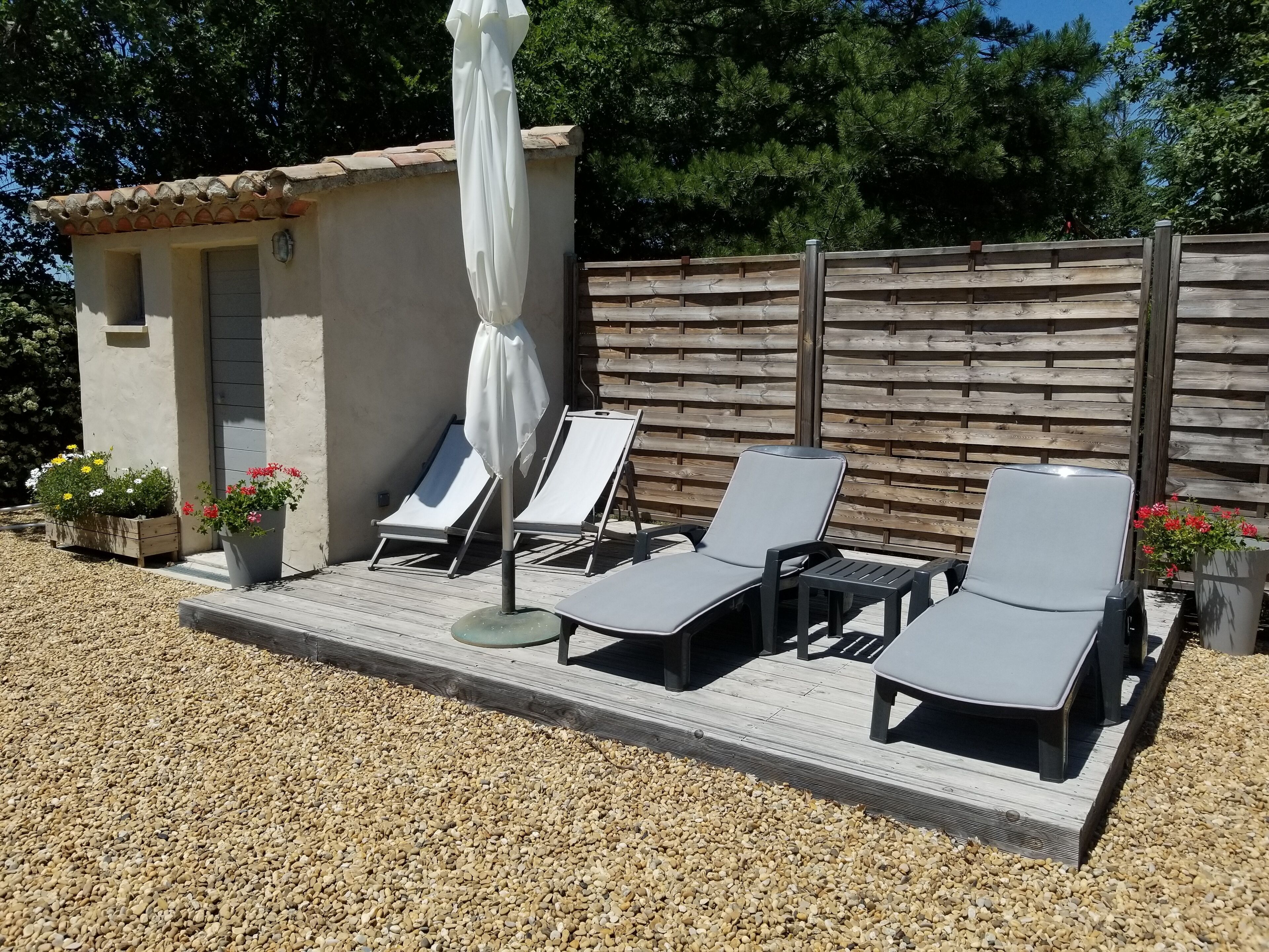 Un espace extérieur ensoleillé, aménagé avec des fauteuils confortables et une petite table, entouré d’une terrasse en bois et d’un sol recouvert de petits galets.