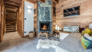 A cozy log cabin interior with a fireplace, a flat-screen TV mounted on the wall, and a comfortable seating area with cushions.