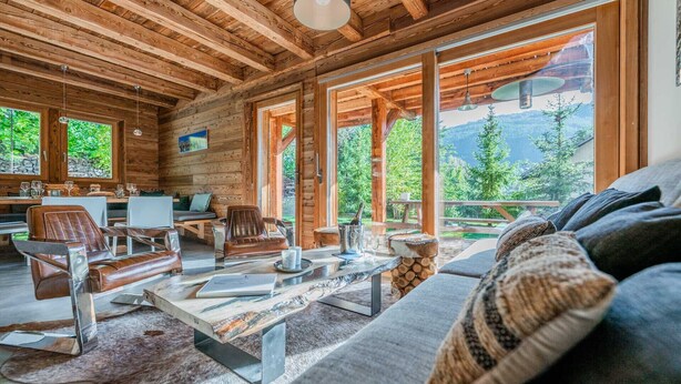 A cozy cabin interior with wooden walls, a dining area, a sofa, and a view of the outdoors.