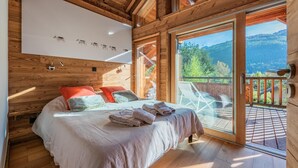 A wooden cabin bedroom with a bed, pillows, and a large window overlooking a deck and mountains.