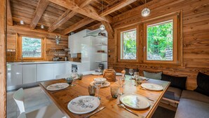 A wooden cabin interior with a dining area, kitchen, and large windows.