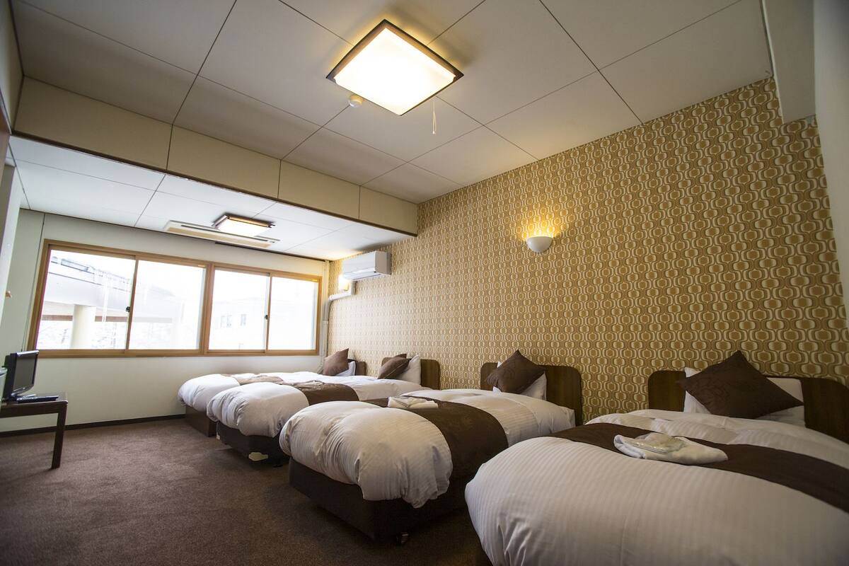 Habitación de hotel con cuatro camas dispuestas en fila, un ventanal con persianas, un televisor de pantalla plana sobre una mesita y un papel tapiz decorado.