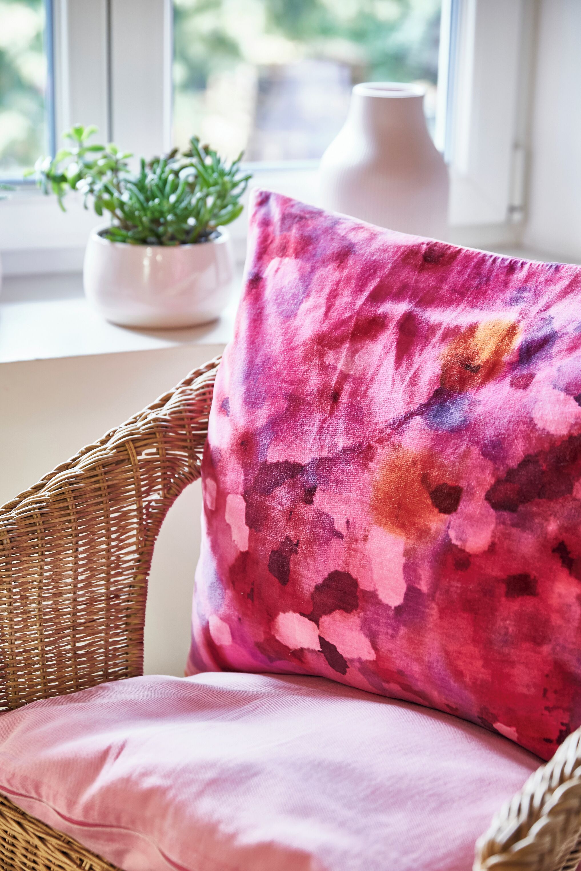 A wicker chair with a pink cushion and a potted plant on a windowsill.