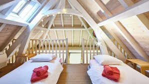 A high-ceilinged attic bedroom with wooden beams, two beds with white linens, and a view through a skylight.