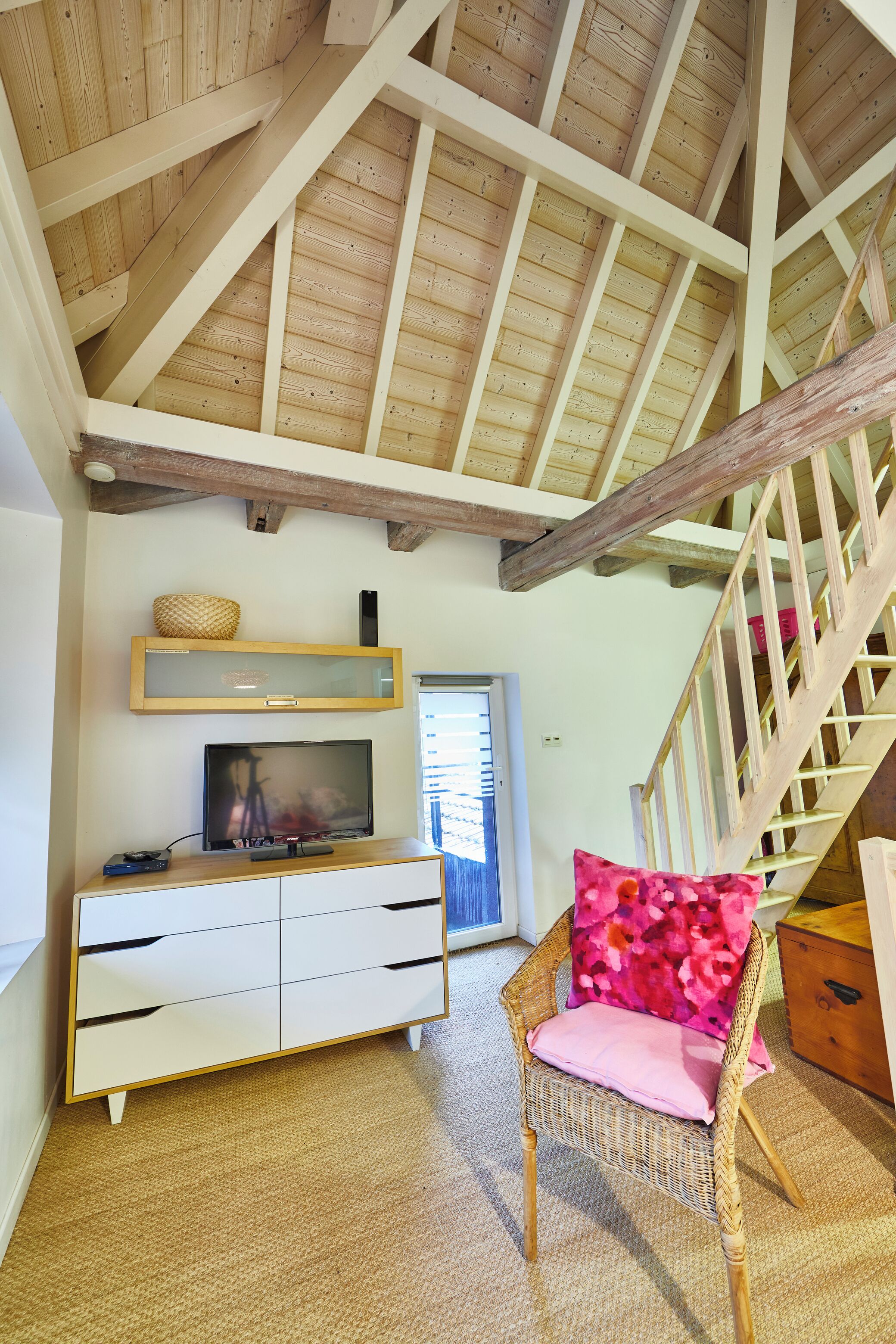 Ein Zimmer mit Holzboden, einer weißen Unterhaltungsecke, einem Korbstuhl mit rosa Kissen und einer Treppe mit Holzwand.